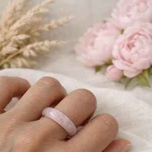 Puede incluir: Un anillo liso y pulido elaborado en piedra de color rosa claro con una apariencia translúcida y veteada. La banda presenta un perfil redondeado y uniforme con un acabado brillante que refleja la luz, mostrando las variaciones naturales.
