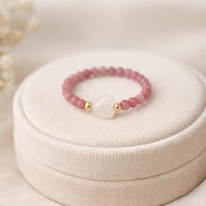 May include: A pink beaded ring with a white heart-shaped stone and gold accents. The ring is set on a white velvet jewelry box. The ring is a delicate piece of jewelry.