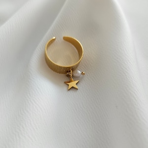 May include: A gold-colored adjustable ring with a textured band. A small gold star and a white pearl dangle from the ring. The ring is set against a white, textured background.