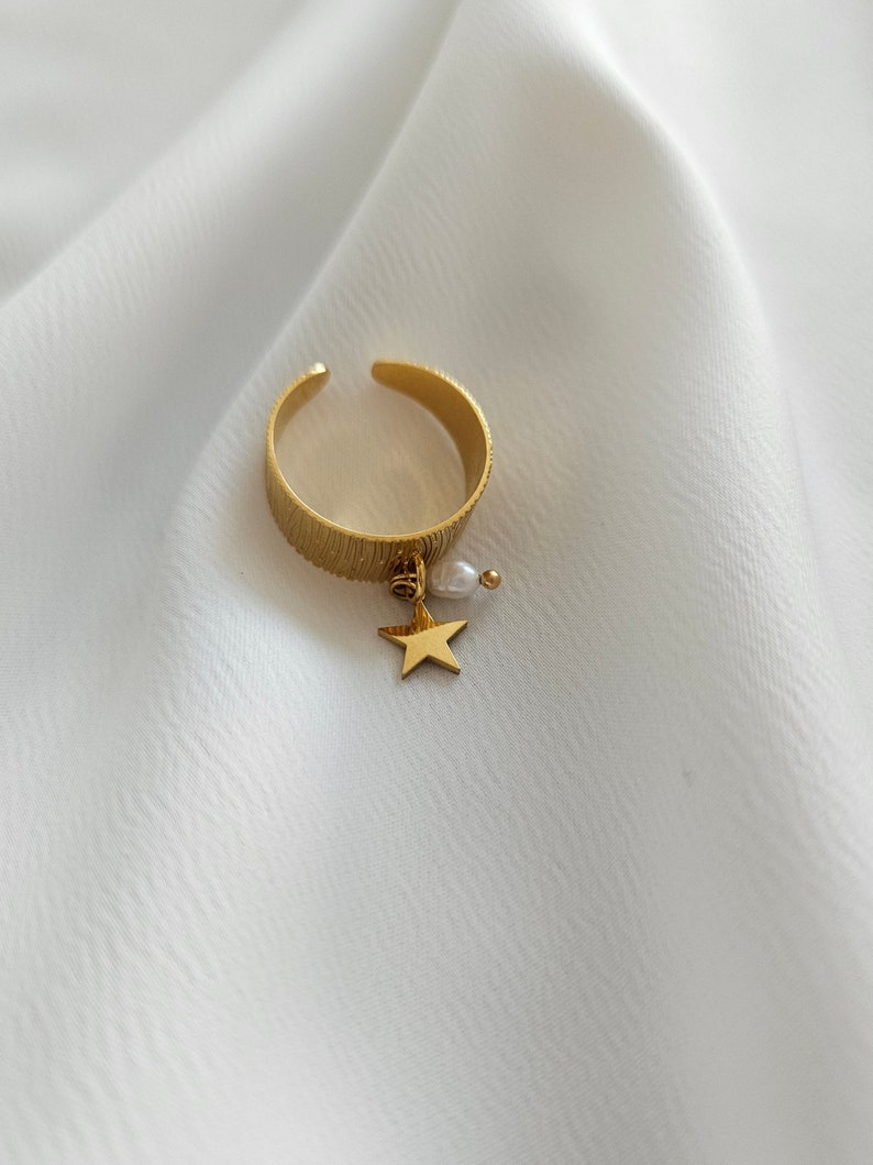 May include: A gold-colored adjustable ring with a textured band. A small gold star and a white pearl dangle from the ring. The ring is set against a white, textured background.