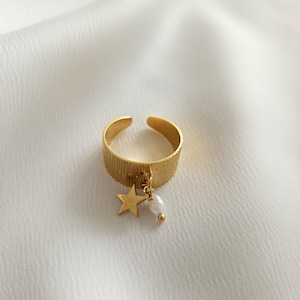 Op de afbeelding: Een goudkleurige verstelbare ring met een getextureerde band. Een kleine gouden ster en een witte parelbedel hangen aan de ring. De ring is geplaatst tegen een witte, getextureerde achtergrond.