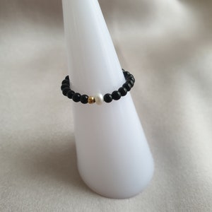 May include: A beaded ring featuring black onyx beads, a single white pearl, and a gold-colored accent bead. The ring is displayed on a white, cone-shaped stand against a soft, neutral background. This piece of jewelry is a stylish accessory.