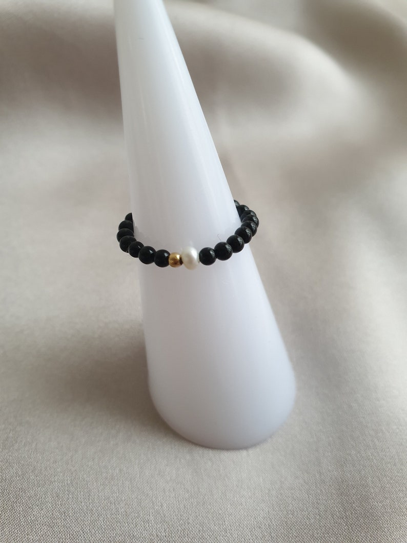 May include: A beaded ring featuring black onyx beads, a single white pearl, and a gold-colored accent bead. The ring is displayed on a white, cone-shaped stand against a soft, neutral background. This piece of jewelry is a stylish accessory.