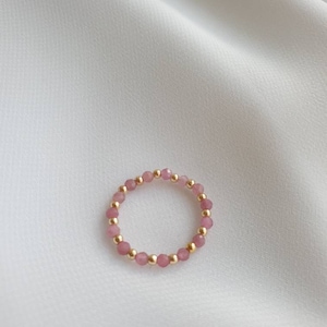 May include: A delicate ring made of small pink and gold beads. The ring is on a white fabric background.