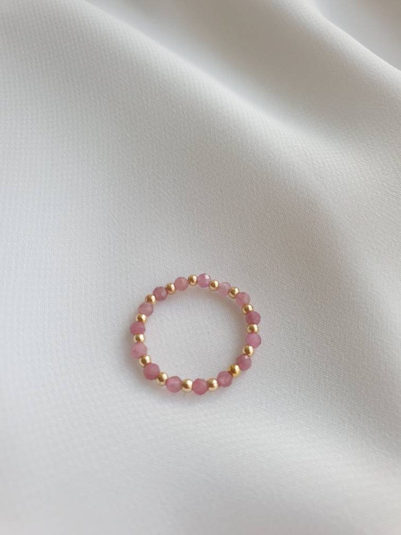 May include: A delicate ring made of small pink and gold beads. The ring is on a white fabric background.