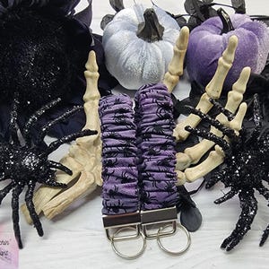 Purple cotton fabric with black spiders design. The keychain uses 7.5 inch to 8 inch wide elastic to make it stretchy. The keychain is completed with silver colored key fob clamps and silver keyring.