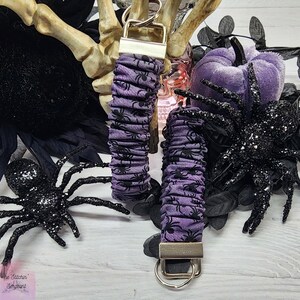 Purple cotton fabric with black spiders design. The keychain uses 7.5 inch to 8 inch wide elastic to make it stretchy. The keychain is completed with silver colored key fob clamps and silver keyring.