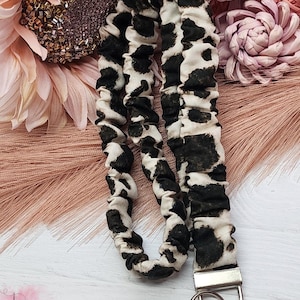 May include: A fabric lanyard featuring a black and white cow print pattern with a gathered, elasticized texture. The lanyard includes a silver-toned metal clasp and a circular split ring at the bottom for attaching keys or identification badges.