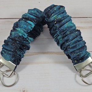 May include: A teal and gold fabric key chain with a silver key ring. The fabric has a galaxy print.