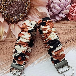 May include: Two fabric wrist keychains with a cow print design in brown, black, and cream colors. Each keychain has a silver-colored metal clasp and a key ring. The keychains are displayed on a light-colored surface.