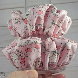 May include: Two pink fabric hair scrunchies with a pig pattern. The scrunchies are made of soft, ruffled fabric and have a gold glitter design.