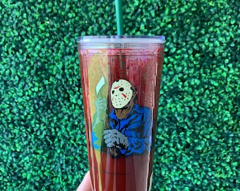 May include: A tall, clear tumbler with a red liquid inside. The tumbler has a graphic of a masked figure holding a weapon. The tumbler has a green straw.