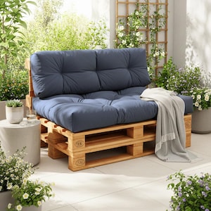 Può includere: Un divano da giardino realizzato con pallet di legno, con un cuscino e schienale blu navy. Una coperta grigio chiaro è drappeggiata sul divano. Il divano è su un patio con piante in vaso e un traliccio.