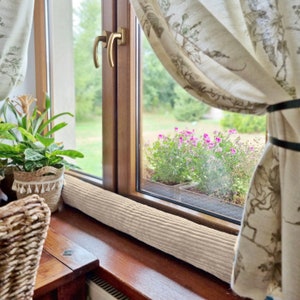 Op de afbeelding: Een beige tochtstopper voor ramen met een geribbelde textuur ligt op een houten vensterbank. Het raam staat open en er is een uitzicht op een tuin met groene planten en roze bloemen te zien door het raam.