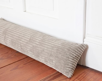 Peut inclure: Un boudin de porte cylindrique et long en tissu velours côtelé beige clair avec une texture côtelée verticale distincte. L'objet est placé horizontalement le long de la base d'un cadre de porte blanc sur un sol en bois.