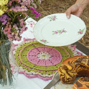Pode incluir: Uma mesa posta com um prato branco com detalhes de rosas cor-de-rosa, uma fatia de bolo mármore e um sousplat de crochê feito à mão em rosa, verde e bege. Um vaso de vidro transparente contém flores coloridas.