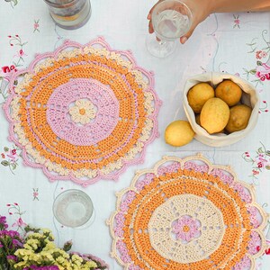 Pode incluir: Duas bases de mesa redondas feitas à mão em crochê, em tons de rosa, laranja e creme, dispostas sobre uma toalha de mesa floral. Uma cesta de limões, um copo de água e uma prensa francesa também são visíveis, sugerindo um ambiente de refeições casual.