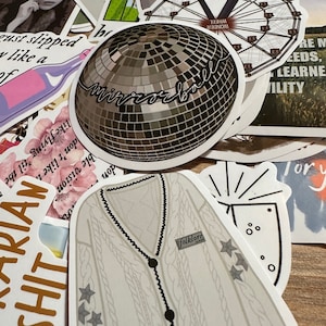 May include: A collection of stickers featuring various designs. Notable stickers include a disco ball with "mirrorball" script, a cable-knit cardigan with "folklore" text, and a Ferris wheel. Other stickers have text and illustrations.