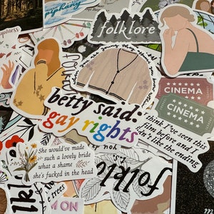 May include: A collection of stickers with various designs and text. Some stickers feature illustrations of a cardigan, a person, and a cinema ticket. Text includes "folklore", "betty said: gay rights", and other phrases.