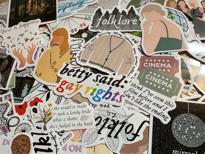May include: A collection of stickers with various designs and text. Some stickers feature illustrations of a cardigan, a person, and a cinema ticket. Text includes "folklore", "betty said: gay rights", and other phrases.