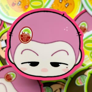 May include: A pink sticker featuring a cartoon face with closed eyes and a small red circle with text. The sticker has a pink border and is surrounded by other stickers with fruit designs.