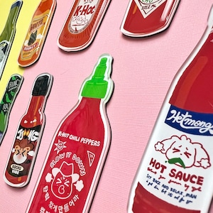 May include: A collection of colorful hot sauce bottles with cartoon illustrations and text. The bottles are red, green, and blue with white lettering. The text on the bottles includes "K-Hot Chili Peppers", "Slow It Down", "Gochu Vibe", "Hetmong", and "Hot Sauce".