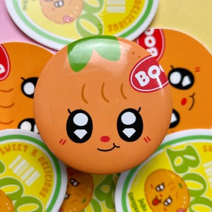 May include: Orange-colored button with a cartoon tangerine face, featuring large black eyes, a small red nose, and a green stem. The button has the text "BQ" on a red sticker. Other buttons with similar designs are in the background.