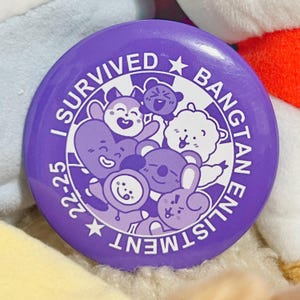 Op de afbeelding: Een paarse knoop met de tekst "I SURVIVED BANGTAN ENLISTMENT 22-25" rondom een groep cartoonfiguren. De knoop heeft een glanzende afwerking en een witte rand. De figuren staan in verschillende poses.