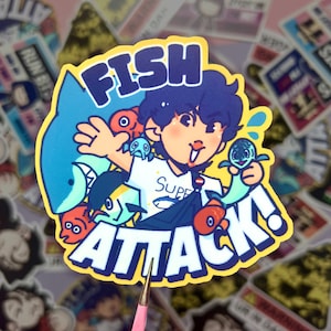 May include: A yellow sticker with a cartoon character wearing a white shirt with the word "SUPER" on it. The character is surrounded by fish, a shark, and a turtle. The sticker says "FISH ATTACK!" in large, bold letters.