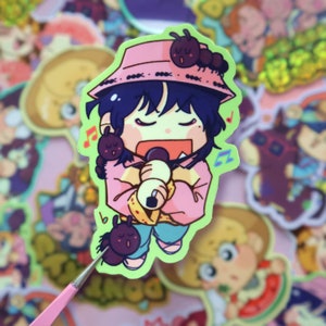 May include: A sticker of a cartoon character singing into a microphone. The character is wearing a pink hat with black accents, a pink jacket, and blue jeans. There are three small black ants on the hat and one on the character's leg. The sticker has a green background.