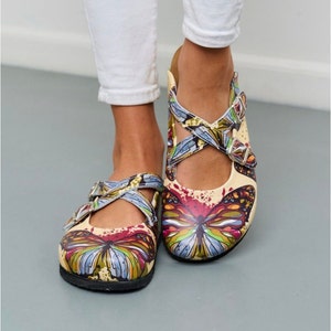 Könnte beinhalten: Ein Paar beigefarbene Slipper mit Schmetterlingsdruck. Die Schuhe haben ein Kreuzriemen-Design mit Schnallenverschluss.