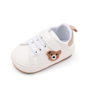 May include: White baby shoes with a brown bear applique and velcro closure.