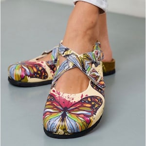 May include: A pair of beige leather sandals with a butterfly print. The sandals have a strap across the foot and a buckle closure. The butterflies are colorful and have a watercolor effect.