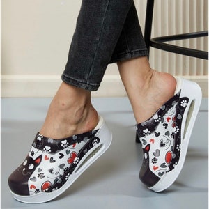 Op de afbeelding: Zwart-witte slip-on klompen met een rood hart- en pootprintontwerp. De klompen hebben een witte platformzool en een zwart-witte kattenafbeelding.