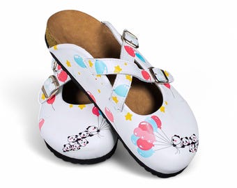 May include: White leather slip-on clogs with a panda and balloon design. The clogs have a cork footbed and a criss-cross strap with a buckle.