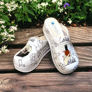 May include: White clogs with a black and white design of hands holding nail polish bottles. The clogs have the word "Nails" written on them.