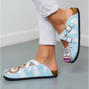 May include: A pair of light blue slip-on shoes with a white strap and an owl design. The shoes have a cork sole and a white cloud pattern.