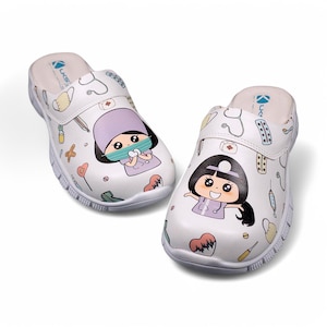 May include: White slip-on clogs with a cartoon nurse design. The clogs have a white background with a colorful cartoon nurse, medical tools, and hearts. The clogs have a white sole.