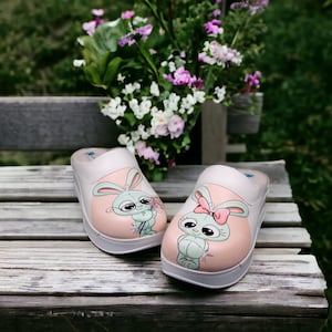May include: A pair of white clogs with a cartoon bunny design. Each clog features a light pink circle with a bunny illustration in the center. One bunny wears glasses and holds flowers, the other has a pink bow. The clogs sit on a wooden surface.