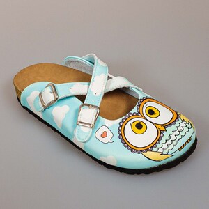 May include: Blue and white slip-on clog with a brown cork sole. The clog has a white strap with a buckle and a graphic of an owl with a heart in a speech bubble.