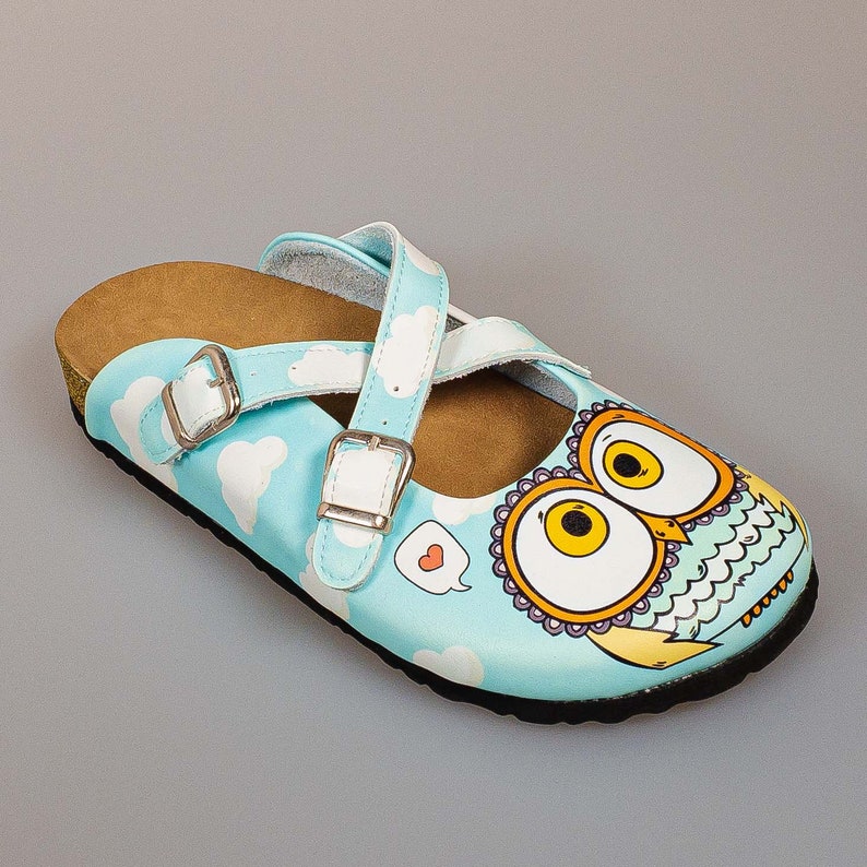 May include: Blue and white slip-on clog with a brown cork sole. The clog has a white strap with a buckle and a graphic of an owl with a heart in a speech bubble.