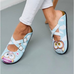 May include: A pair of blue and white slip-on shoes with an owl design. The shoes have a white strap with buckles across the top.