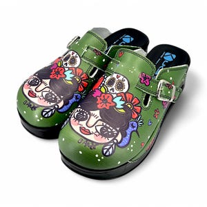 May include: A pair of green clogs with a colorful floral and skull design. The clogs have a black strap with a buckle and a white polka dot pattern.