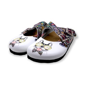 May include: White leather clogs with a colorful, abstract pattern and a cartoon dog wearing sunglasses and a bow tie on the front.