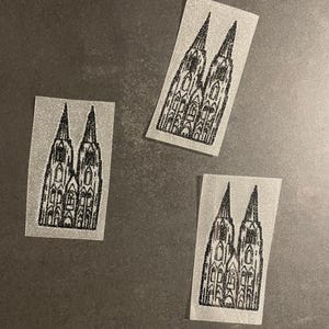 Könnte beinhalten: Drei Aufkleber mit einer schwarz-weißen Darstellung des Kölner Doms. Die Aufkleber sind rechteckig mit einem glitzernden, silbergrauen Hintergrund. Das Dom-Design ist mittig platziert.