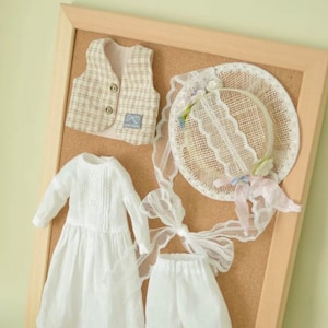K&ouml;nnte beinhalten: Ein Set Puppenkleidung, das auf einer Korktafel ausgestellt ist. Das Set beinhaltet ein wei&szlig;es Kleid mit Spitzenbesatz, eine wei&szlig;e Hose mit Spitzenbesatz, eine karierte Weste und einen Strohhut mit Spitzen- und Blumenapplikationen.