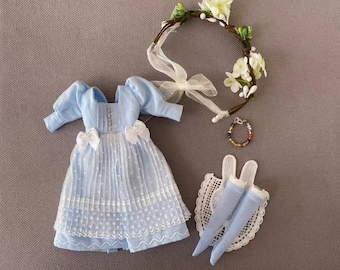 May include: A light blue dress with white lace trim and a white bow on the bodice. The dress is for a doll and is accompanied by a white crocheted bonnet, a pair of blue tights, and a flower crown.