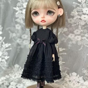 May include: A black doll with blonde hair wearing a black dress with ruffles and lace. The doll has large blue eyes and is wearing black boots.