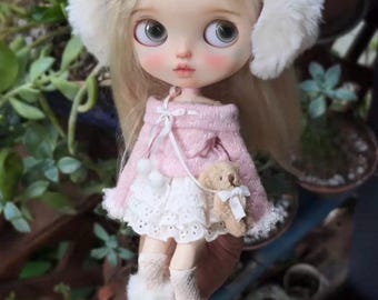 Può includere: Una bambola Blythe personalizzata con capelli biondi, che indossa un maglione rosa, una gonna di pizzo bianco e stivali bianchi soffici. La bambola ha un fiocco rosa, paraorecchie e un piccolo accessorio orsetto. La bambola è tenuta in mano.