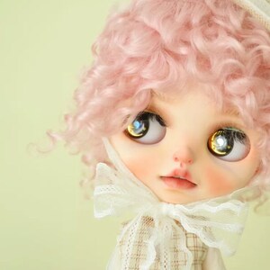 K&ouml;nnte beinhalten: Eine Puppe mit pinkfarbenen, lockigen Haaren, die einen wei&szlig;en Hut und eine wei&szlig;e Schleife aus Spitze tr&auml;gt. Die Puppe hat gro&szlig;e, blaue Augen mit goldenen Flecken.